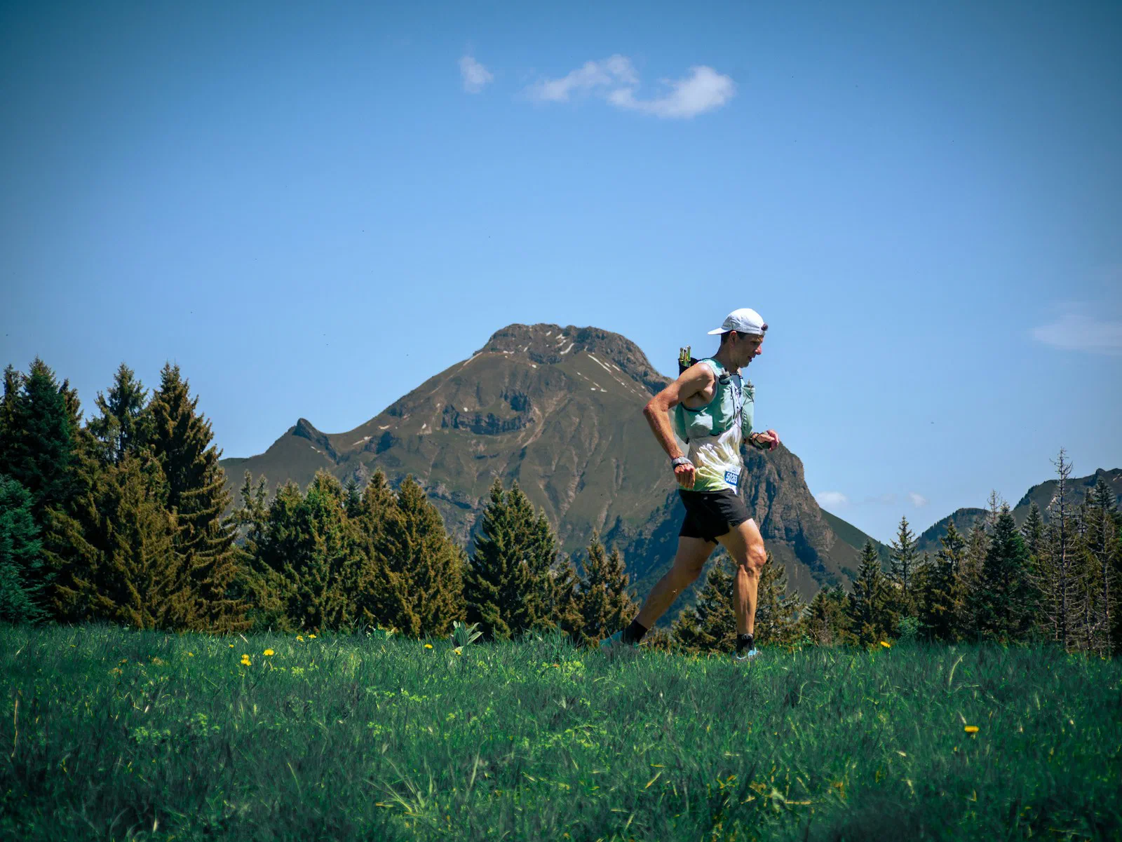 A trail runner crossing a mountain meadow with a rocky peak behind, mid-stride in bright daylight